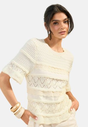 Jonge vrouw met donker haar, gekleed in een offwhite gestructureerd gebreid kortemouw topje en crèmekleurige armbanden, poserend met handen in de zakken.