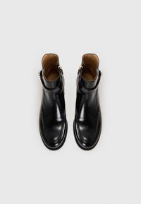 Botas de tornozelo em couro preto com uma textura suave, bico arredondado e detalhe em zíper dourado. O interior apresenta um forro em tom bege para contraste.
