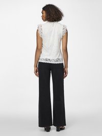 Femeie cu păr creț, văzută din spate, purtând un top alb din dantelă fără mâneci și pantaloni negri largi, cu pantofi cu toc negri.