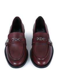 Mocasines de cuero burdeos con punta redondeada, detalle decorativo de cadena plateada y suela de goma negra. Textura suave con un brillo sutil.