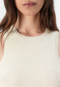 Hladký bezrukávový top v světle béžové barvě, s kulatým výstřihem a měkkou, strukturovanou tkaninou, která těsně přiléhá k tělu.