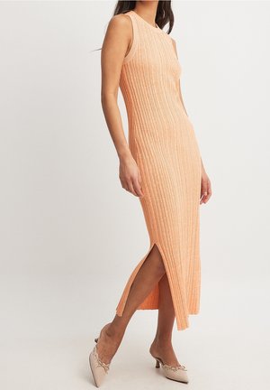 Ærmeløs, ribbet ferskenfarvet midi-kjole med sideslids båret af en kvinde kombineret med beige kitten heel-sko mod ensfarvet baggrund.