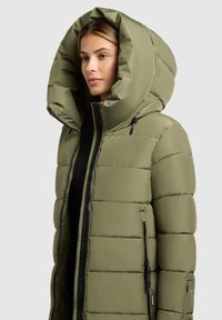 Olivengrüner Puffer-Mantel mit großer Kapuze, ausgestattet mit einem schwarzen Reißverschluss und seitlichen Reißverschlusstaschen. Glatte Textur, gestepptes Design.