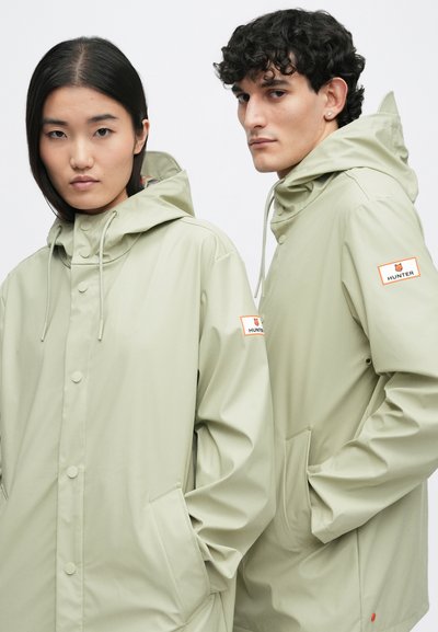 Dwie modelki ubrane w jasnozielone kurtki przeciwdeszczowe z kapturem, każda z naszywką z logo Hunter na lewym rękawie, pozują na jednolitym tle.