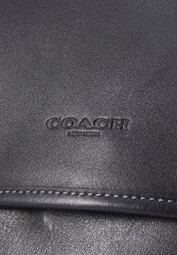 Juodas odinis audinys su tekstūruota danga. Pavaizduotas iškilus "COACH" logotipas su "NEW YORK" po juo. Matoma siūlė palei kraštą.