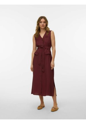 Nő áll, ujjatlan, bordó, gombos középhosszú ruhát visel, melynek derekán kötött öv van, és barna lapos cipőt visel, fehér háttér előtt.