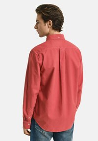Rotes Hemd mit Knöpfen und einem Rückenfalten, langen Ärmeln und geknöpften Manschetten. Getragen mit blauen Jeans. Glatte Textur, lässiges Design.
