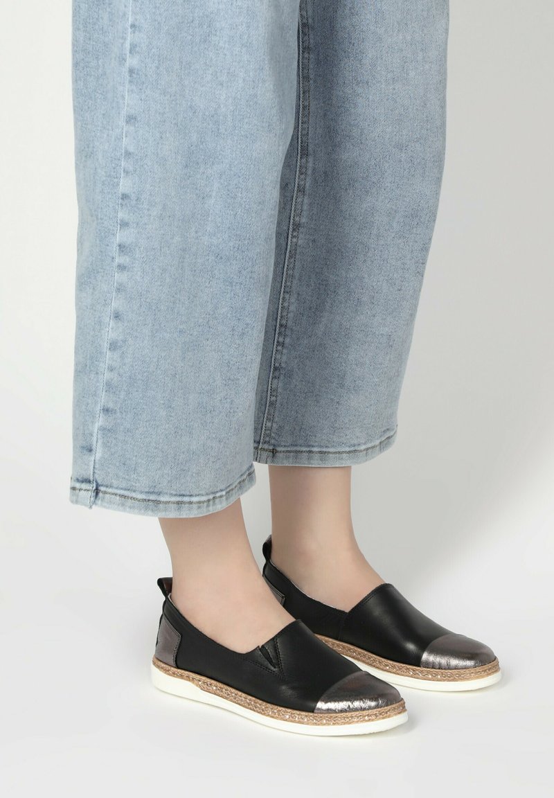 Černé slip-on boty s metalickou špičkou a tkaným detailem podél podrážky, spárované se světle modrými zkrácenými denimovými kalhotami.
