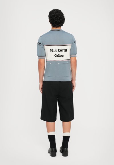 Top de punto azul claro, de manga corta, con detalles en blanco y negro, que presenta el logo "Paul Smith Ciclismo", combinado con pantalones cortos y calcetines negros.