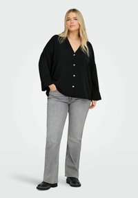 Črna srajca z gumbki in V-izrezom, ohlapen kroj in dolge rokave, v kombinaciji s sivimi ravnimi hlačami in črnimi močnimi škornji.