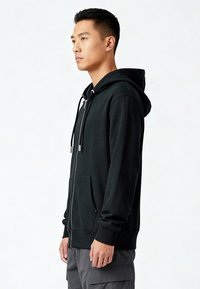 Giovane uomo in profilo che indossa una felpa nera con zip e cappuccio e pantaloni cargo grigi, su uno sfondo bianco semplice.