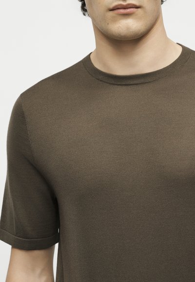 Νέος άνδρας που φοράει μια μονόχρωμη ελαφρώς βαμμένη ελαφριά ελιάς κοντομάνικη πλεκτή μπλούζα με στρογγυλή λαιμόκοψη, κομμένη κάτω από τη μύτη.