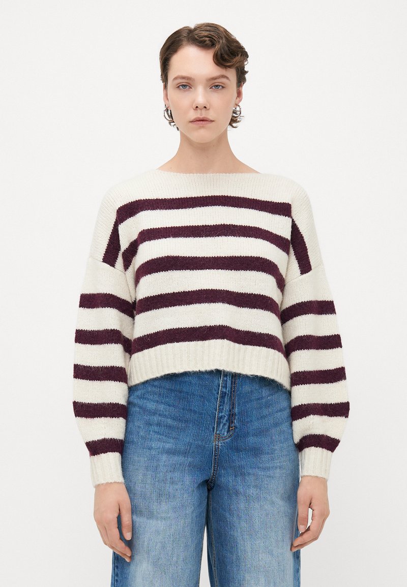 Frau mit kurzen, lockigen Haaren, die große Ohrringe, einen gestrickten Pullover in Weiß und dunkelrot gestreift sowie blaue High-Waist-Jeans trägt und vor einem weißen Hintergrund steht.