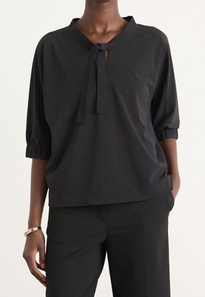 Person trägt ein schwarzes Langarmoberteil mit vorderer Schleifendetail und schwarze Hose, mit einer Hand in der Tasche und einem goldenen Armband am Handgelenk.