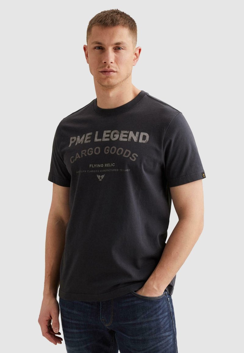 Jeune homme aux cheveux courts portant un T-shirt noir PME Legend Cargo Goods et un jean bleu foncé, debout avec une main dans la poche.