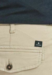 Beżowe bawełniane spodnie z zapięciem na guzik i kieszenią naszywaną. Posiadają czarną metkę z białym napisem "JACK & JONES."