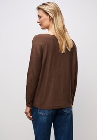 Maglione morbido a maglia marrone con vestibilità rilassata e scollo ampio, abbinato a jeans in denim blu. La texture morbida è visibile nel tessuto.