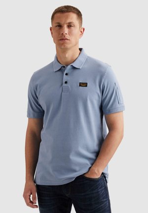Mann trägt ein hellblaues kurzärmeliges Poloshirt mit Kragen, Knopfleiste, Brusttasche und Tasche am linken Ärmel, kombiniert mit dunkler Jeans.