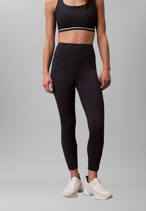 Femme portant un soutien-gorge de sport noir, un legging noir taille haute et des baskets blanches épaisses, debout devant un fond gris uni.
