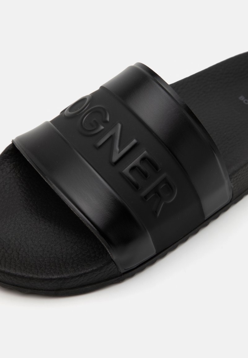 Schwarze Gummisandale mit strukturiertem Fußbett und einem breiten Riemen, der mit dem Markennamen geprägt ist. Flache Sohle mit subtilen Gripp-Details.