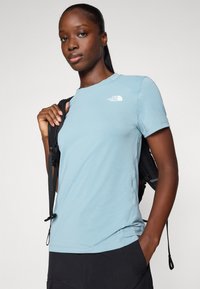 Lichtblauw shirt met korte mouwen van zacht stof, met een wit logo op de bovenste linkerzijde. Model houdt een zwarte rugzakriem vast.