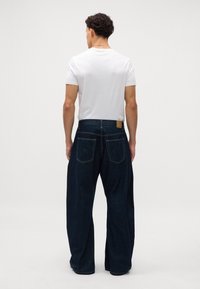 Pantalones de mezclilla de pierna ancha en azul oscuro con costuras claras y parches en la cintura, combinados con una camiseta básica blanca de cuello redondo.