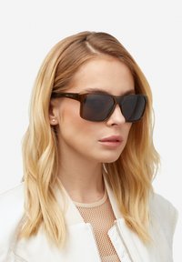 Pruunid, ruudukujulised päikeseprillid tumedate toonitud läätsedega. Valmistatud läikivast materjalist, kombineeritud helevärvilise bomberjope ja võrktõrvikuga.