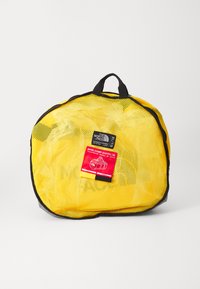 Bolsa de deporte de malla amarilla con ribetes negros, que cuenta con un asa superior y una etiqueta roja que muestra los detalles del producto y sus dimensiones.