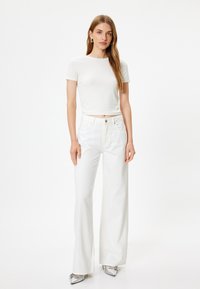 Hvid cropped T-shirt parret med højtaljede hvide flare jeans. Modellen har metallic sølv sko på. Glat stof, rene linjer, minimalistisk design.