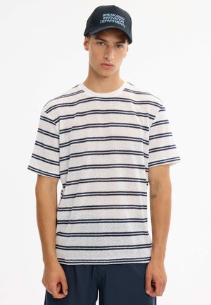 Joven con una camisa blanca de manga corta con rayas horizontales azul marino y una gorra negra con texto blanco, de pie frente a un fondo liso.