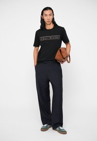 Mann med svart grafisk t-skjorte, stripete vid bukse, grønne sneakers, holder en brettet brun pose med den ene hånden.