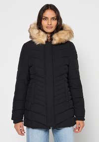 Manteau matelassé noir avec capuche en fausse fourrure, présentant un design matelassé en chevrons et des poches latérales. Texture lisse, coupe mi-longue.