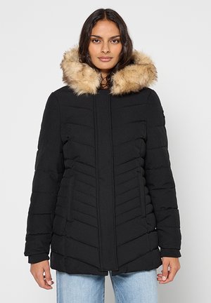 Manteau matelassé noir avec capuche en fausse fourrure, présentant un design matelassé en chevrons et des poches latérales. Texture lisse, coupe mi-longue.