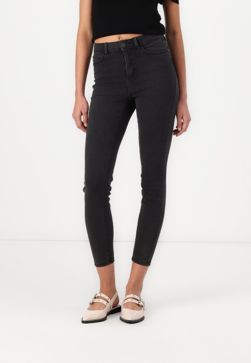 Femme portant un jean skinny noir taille haute coupé au-dessus de la cheville avec des ballerines crème pointues à double brides et boucles.