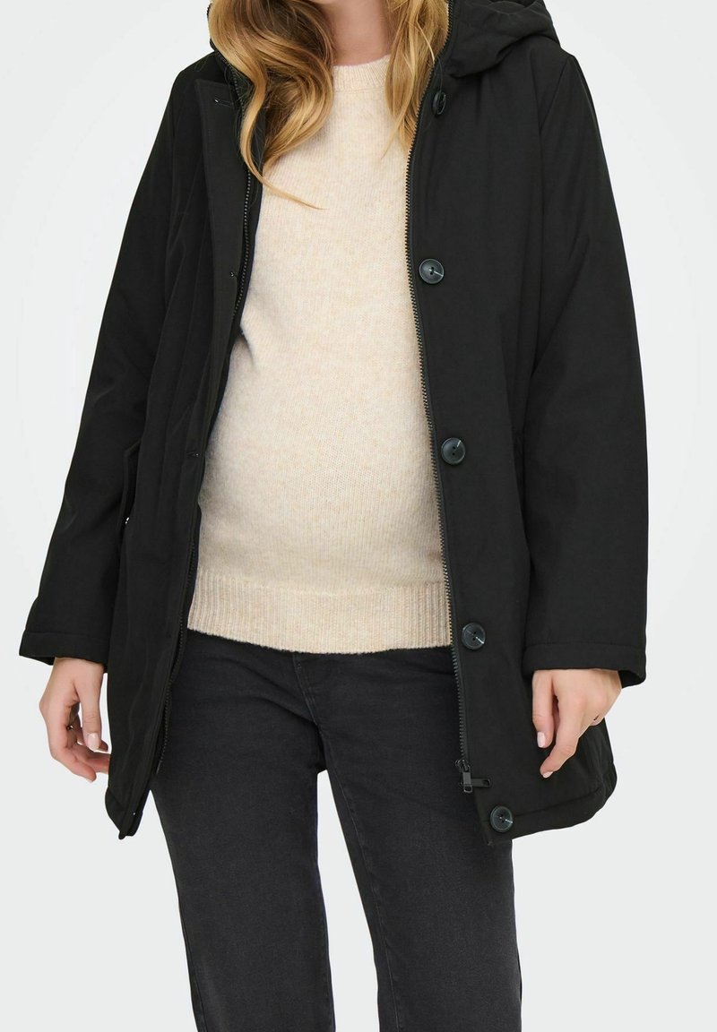 Femme portant un manteau noir à capuche par-dessus un pull en maille crème et un pantalon noir, debout devant un fond clair uni.