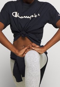 Camiseta de algodón navy con un frente anudado y el logo blanco de "Champion", combinada con leggings gris claro; textura suave y ajuste relajado.
