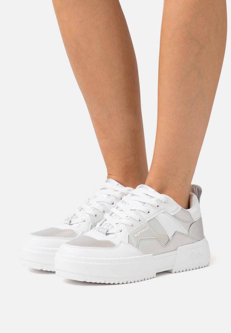 Zapatilla blanca con acentos grises; presenta una suela gruesa, diseño de cordones y parte superior sintética con una textura suave y un detalle de logo.