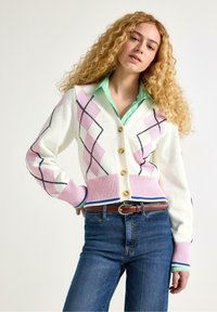 Pletený bílý kardigan s růžovo-černými vzory argyle, s výstřihem ve tvaru V, zapínáním na knoflíky a žebrovaným lemem. Kombinován se zeleným pruhovaným tričkem.
