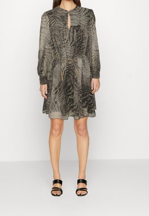 Femme portant une robe longue aux genoux à manches longues avec motif abstrait noir et gris, ceinture cintrée à la taille et talons ouverts noirs, debout sur fond uni.
