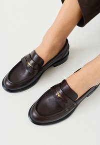 Brown leather loafers with a smooth texture, raised stitching, a strap, and metallic accent; flat sole and rounded toe design.