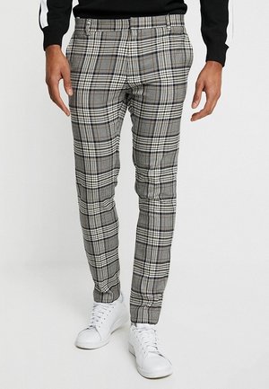 Man draagt een slim-fit grijze geruite broek gecombineerd met witte sneakers en een zwarte trui, staand tegen een witte achtergrond.