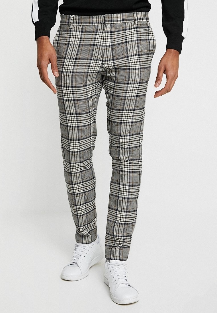 Homme portant un pantalon slim gris à carreaux associé à des baskets blanches et un pull noir, debout devant un fond blanc.