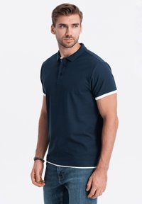 Námořnické modré polo tričko s černými knoflíky, bílými akcenty na límci a krátkými rukávy, v kombinaci se světle modrými džíny. Hladká bavlněná látka.