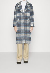 Vlněný kabát s kostičkovaným vzorem v modrých, krémových a hnědých tónech. Má klopový límec s výstřihem, dlouhé rukávy, dvě přední kapsy a zapínání na knoflíky.