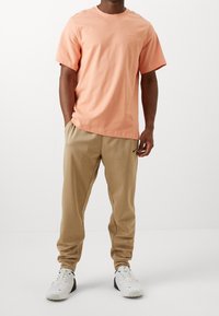 Camiseta de algodón rosa claro con mangas cortas y cuello redondo, combinada con pantalones jogger beige que tienen un logo negro, llevados con zapatillas blancas.