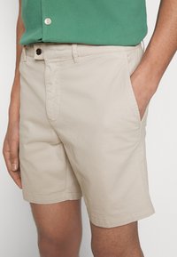 Beige bomullsshorts med rak passform. Har sidfickor, knappstängning och en slät yta. Bärs med en grön skjorta.