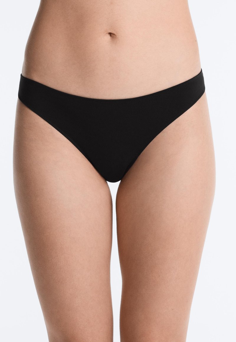 Femme portant une lingerie noire style bikini sans coutures, montrée de la taille à mi-cuisse sur fond blanc.