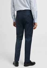Pantalons ajustés bleu marine avec une texture lisse, devant plat et deux poches arrière. Portés avec une chemise à manches longues bleu clair et des chaussures noires.