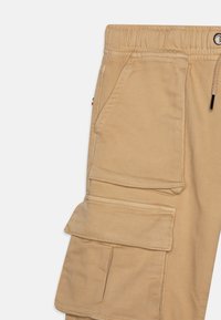 Beige Cargo-Hosen mit elastischem Taillenbund, Kordelzug und mehreren aufgesetzten Taschen mit Steppdetails.