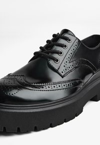 Scarpe nere in pelle con dettagli brogue, caratterizzate da perforazioni, design con lacci e una suola in gomma spessa e rigata. Finitura liscia e lucida.